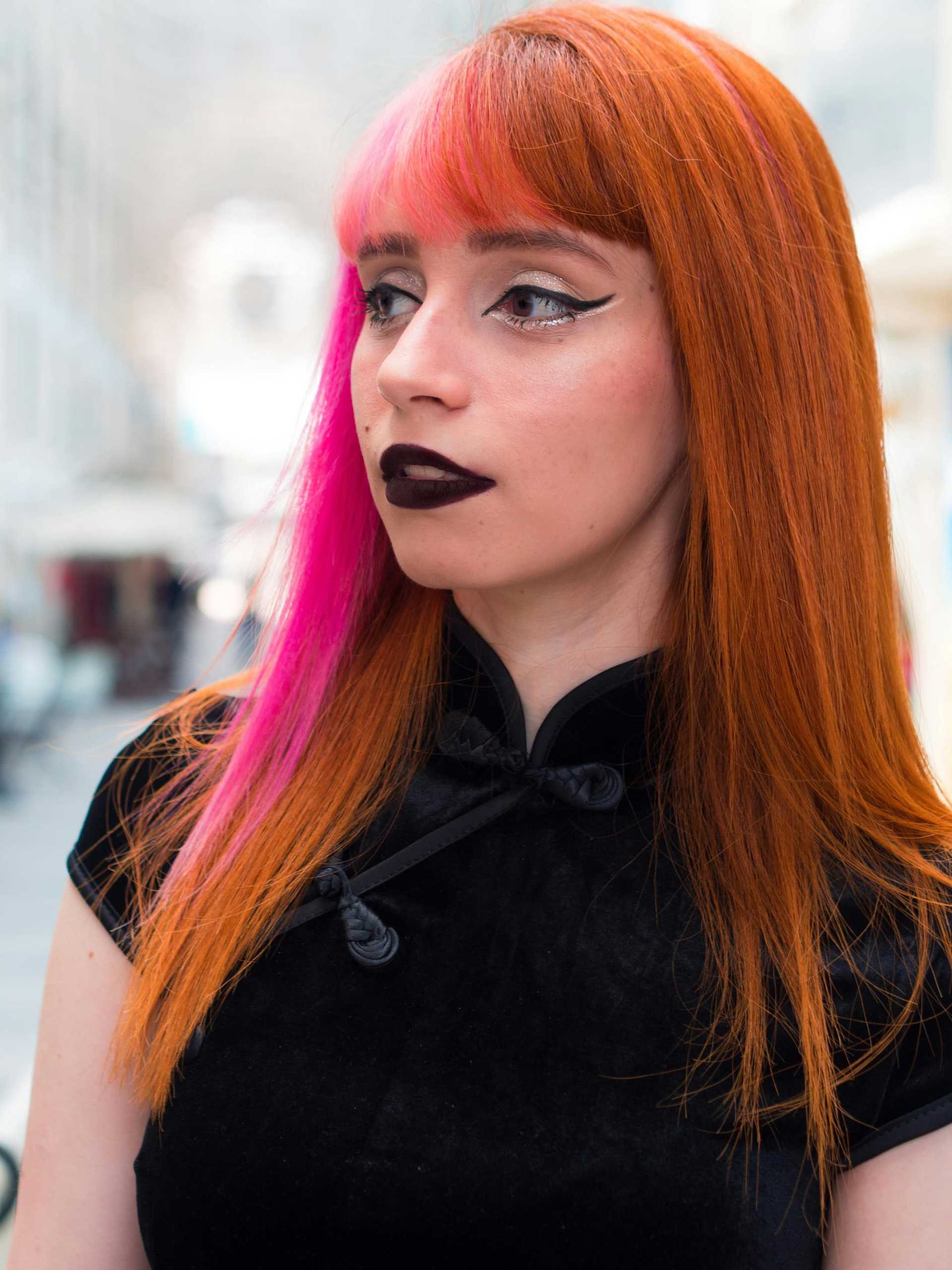 Woman with orange and pink hair, wearing black lipstick and a dark dress in a city street.