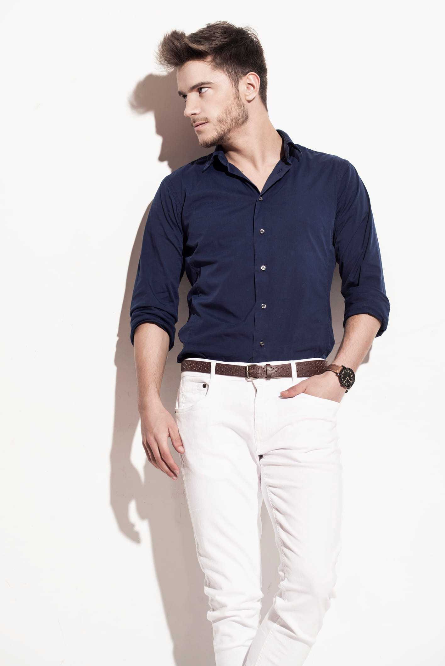 Man in blue shirt and white pants, standing against a white background, looking to the side.