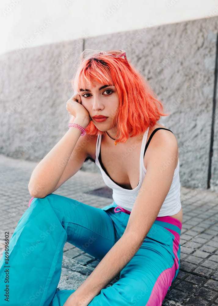 Young person with bright orange hair sitting on pavement in casual attire.